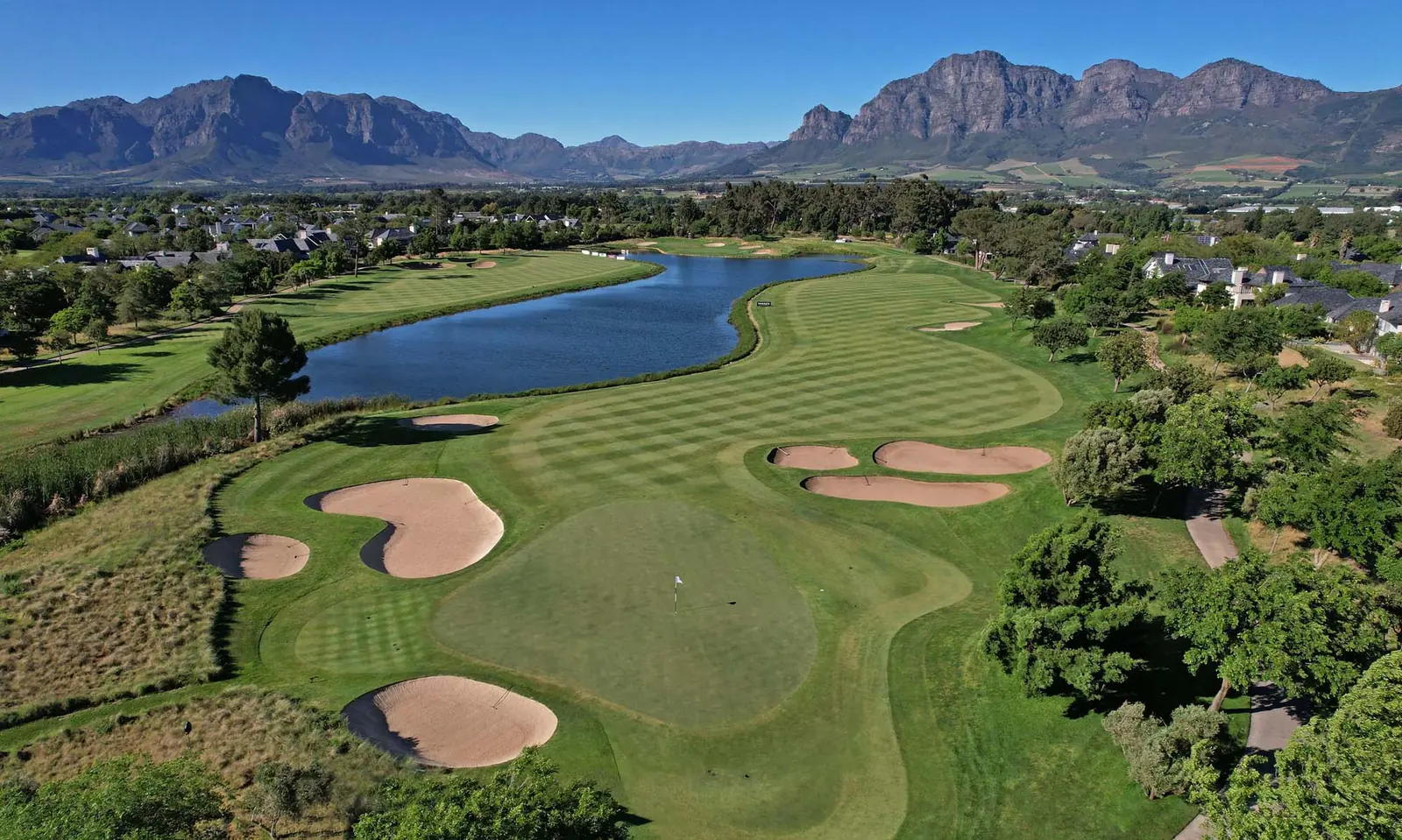 Aerial view of a golf hole with striped fairways, bunkers and a lake, set against mountains under a clear blue sky